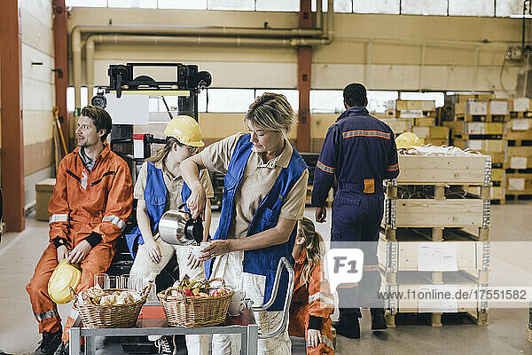 Weiblicher Arbeiter gießt Kaffee in eine Tasse  während Kollegen im Hintergrund in einem Fabriklager zu sehen sind