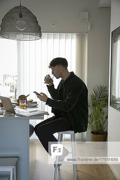 Junger Geschäftsmann trinkt aus einem Glas  während er zu Hause sein Smartphone benutzt