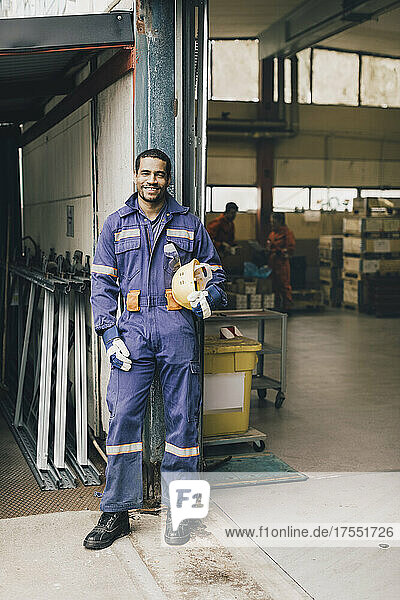 Porträt eines lächelnden männlichen Arbeiters in Arbeitsschutzkleidung am Eingang einer Fabrik