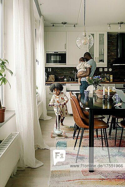Vater trägt kleinen Jungen  während seine Tochter in der Küche Roller fährt
