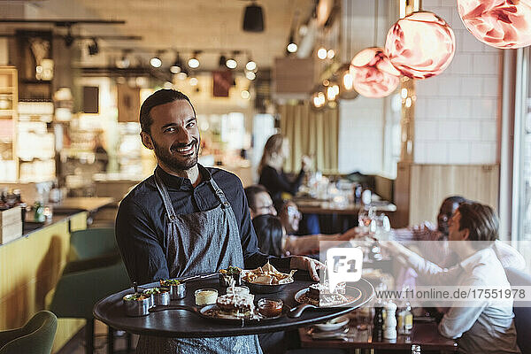 Porträt eines männlichen Kellners mit einem Essenstablett  während die Kunden im Hintergrund an der Bar anstoßen