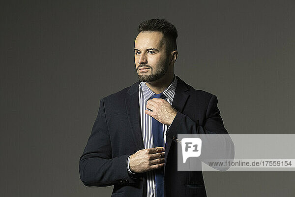 Portrait confident  stylish businessman in suit adjusting tie against black background
