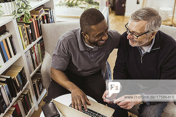 Happy male caregiver teaching online shopping to senior man on laptop at nursing home