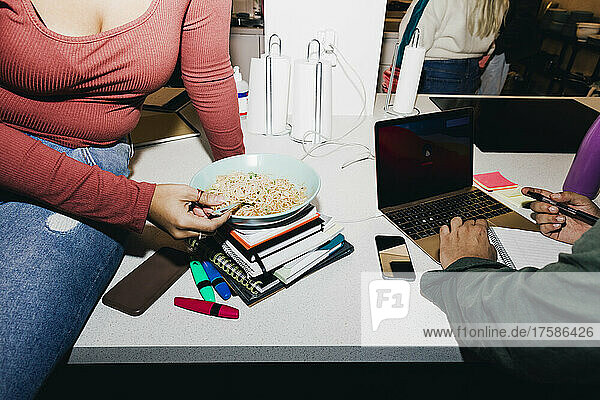 Midsection woman having noodles while male friend using laptop in college dorm