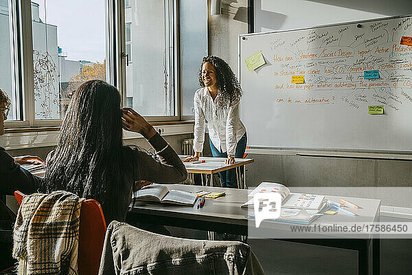Fröhlicher Professor unterrichtet Universitätsstudenten  während er sich auf den Schreibtisch im Klassenzimmer stützt