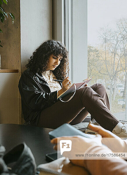 Young woman writing in book while sitting by window at university