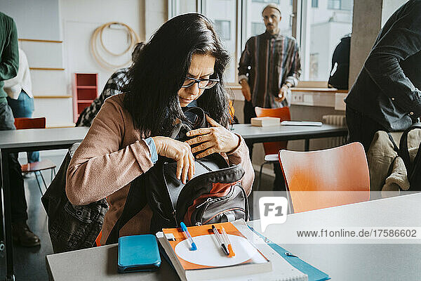 Reife Studentin nimmt Bücher aus dem Rucksack  während sie am Schreibtisch im Klassenzimmer sitzt