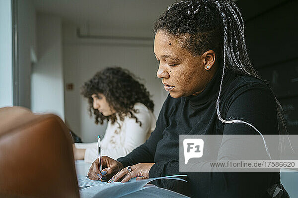 Woman with braided hair writing in book while sitting by friend at university