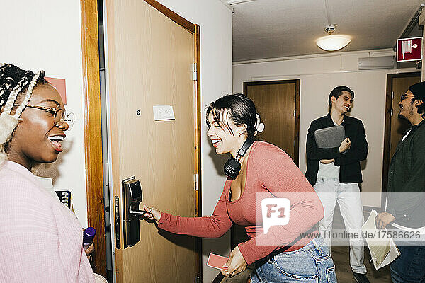 Glückliche junge Frau  die die Tür öffnet  während ihre multirassischen Freunde auf dem Flur des Studentenwohnheims stehen