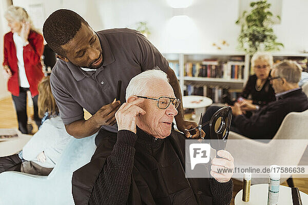 Male healthcare worker talking with senior man while cutting hair in retirement home