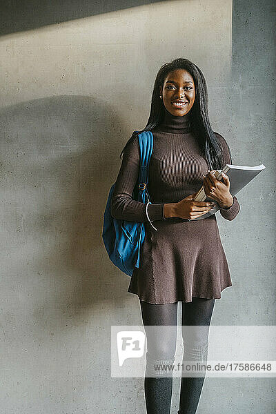 Ganzkörperporträt einer lächelnden jungen Frau  die ein Buch in der Hand hält und mit einem Rucksack an einer grauen Wand steht