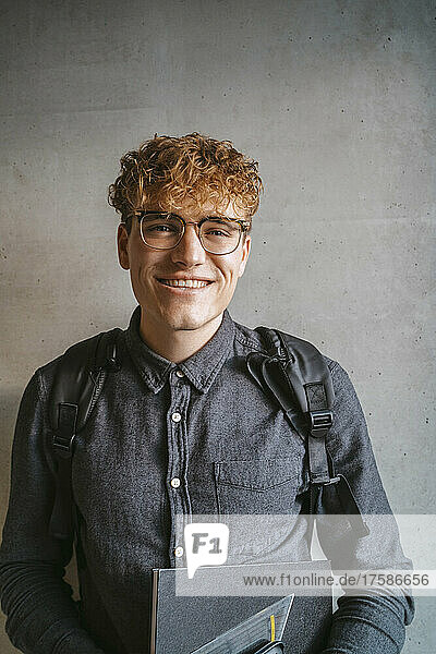 Porträt eines lächelnden blonden männlichen Studenten mit Brille  der ein Buch gegen eine graue Wand hält