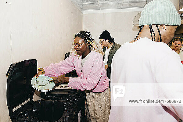 Junge Frau kratzt Essensreste in den Mülleimer  während Freunde in der Küche des Studentenwohnheims sitzen