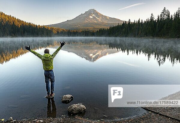 Junger Mann steht auf Stein und streckt die Arme in die Luft  Spiegelung des Vulkans Mt. Hood im See Trillium Lake  bei Sonnenaufgang  Oregon  USA  Nordamerika
