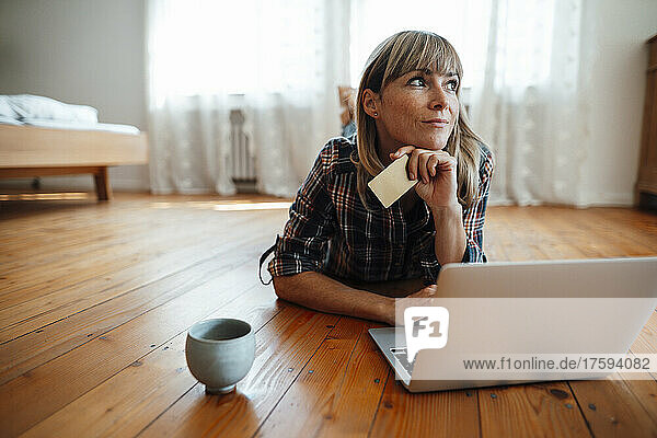 Nachdenkliche Frau mit Kreditkarte und Laptop  die zu Hause auf dem Boden liegt