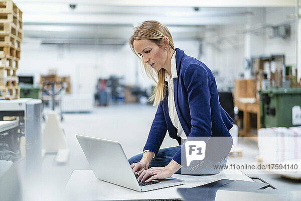 Businesswoman using laptop sitting on desk in factory