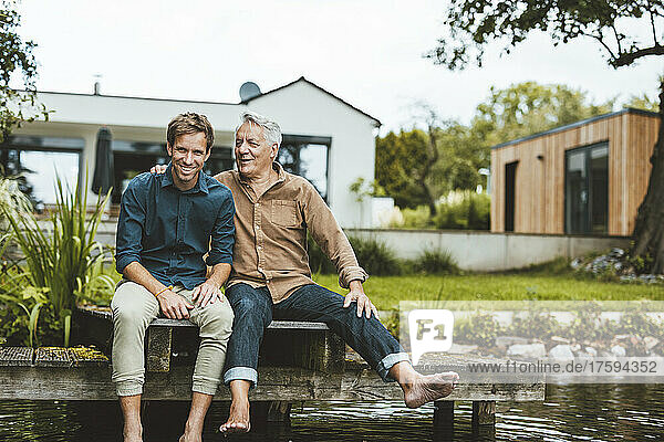 Happy father and son sitting on jetty by lake at backyard