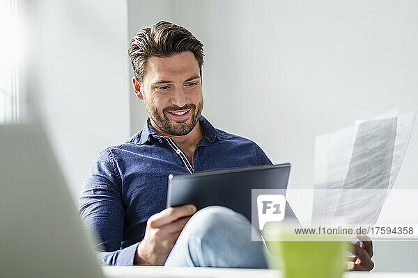 Smiling businessman with document working on tablet PC in office