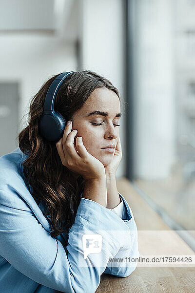 Young businesswoman with head in hands listening music through wireless headphones at desk