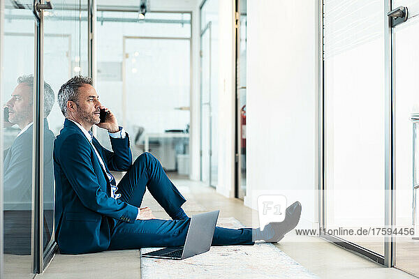 Thoughtful businessman sitting with laptop talking on mobile phone in office corridor