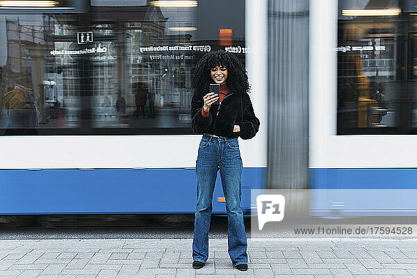 Fashionable young woman using smart phone in front of moving tram