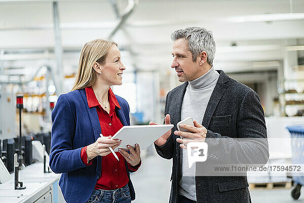 Smiling blond businesswoman with tablet PC looking at colleague discussing ideas in factory
