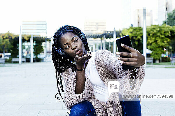 Young woman wearing headphones crouching while blowing kiss on smart phone