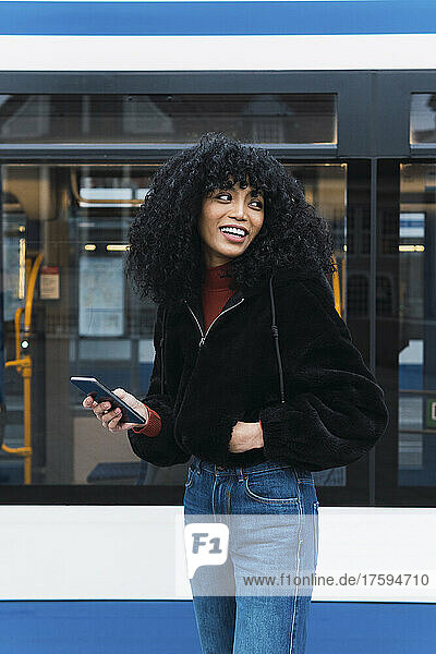 Smiling young woman with smart phone in front of tram
