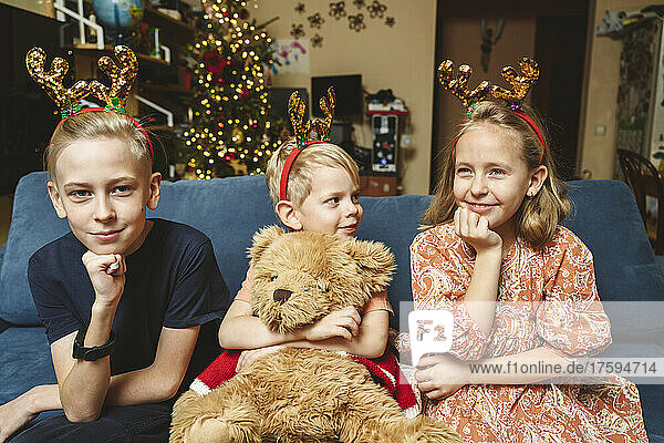 Siblings wearing reindeer antler sitting on sofa at Christmas