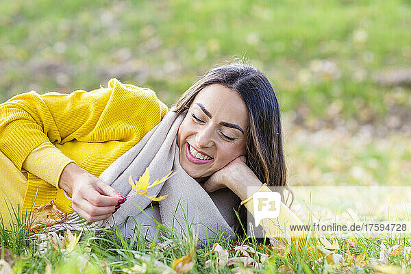 Happy young woman with leaf lying on grass in autumn park