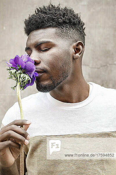 Young man with eyes closed smelling flower in front of wall