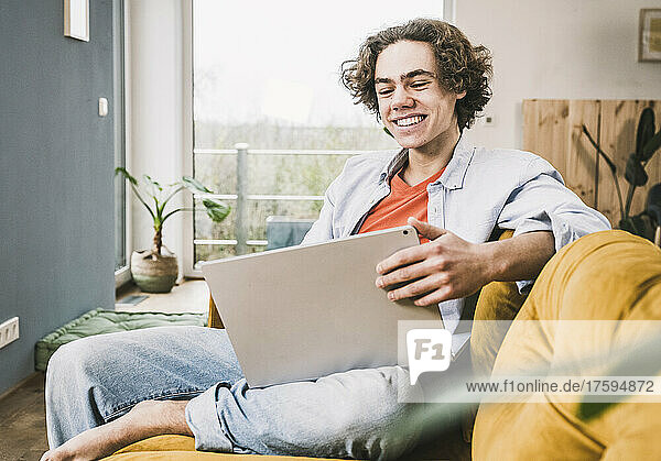 Glücklicher junger Mann sitzt mit Laptop auf dem Sofa im Wohnzimmer