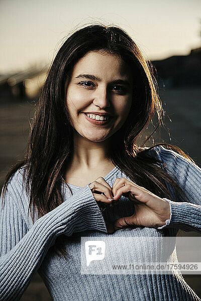 Smiling young woman making heart shape with hands at sunset