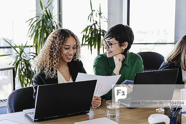 Happy businesswomen reviewing financial report with laptops at desk in coworking office