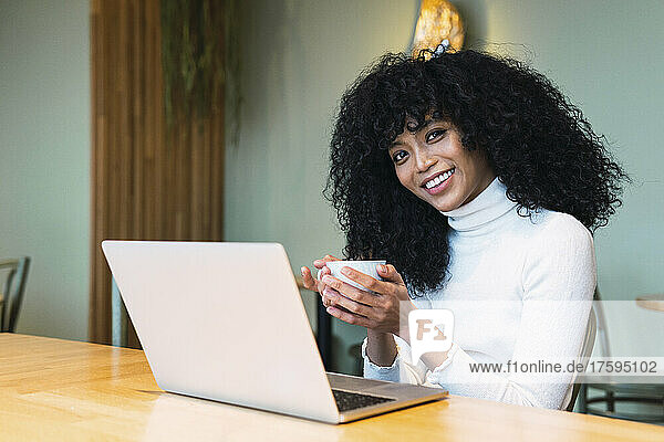 Smiling curly black haired woman holding coffee cup sitting with laptop at cafe