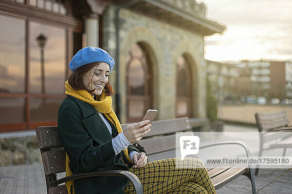 Smiling woman surfing net through mobile phone sitting on bench