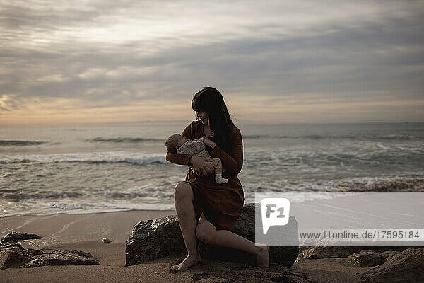 Caring mother holding newborn son sitting on rock at beach