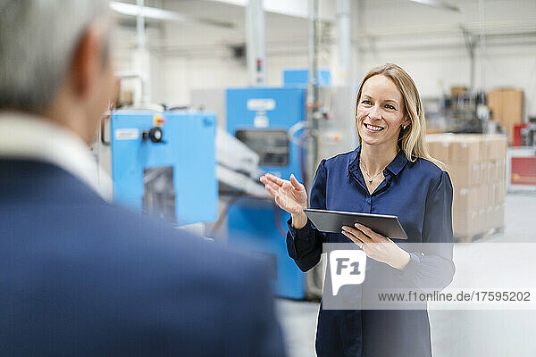 Smiling blond businesswoman holding tablet PC discussing with colleague in factory