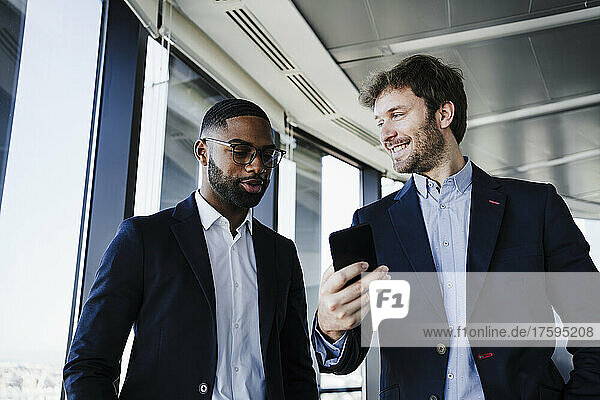 Smiling businessman with smart phone looking at colleague talking in office
