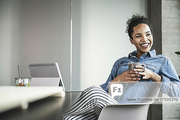 Happy businesswoman with coffee cup sitting on chair in office