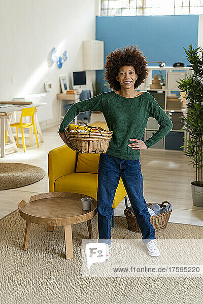 Smiling Afro woman with hand on hip holding basket in living room
