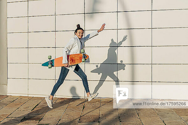 Cheerful woman jumping with skateboard on footpath