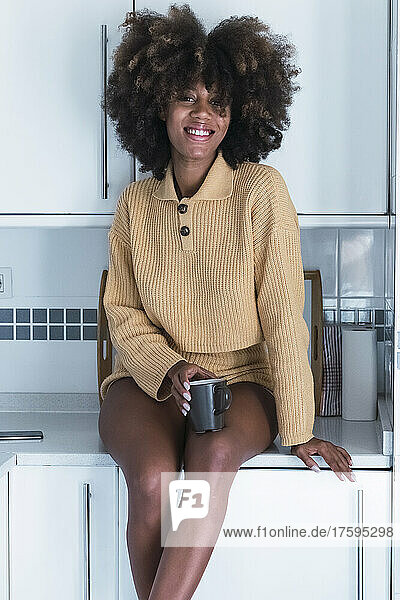 Young woman with Afro hairstyle sitting on counter in kitchen