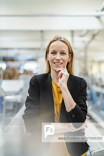 Happy blond businesswoman standing with hand on chin in factory