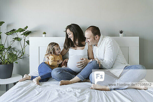 Happy man and woman looking at daughter sitting on bed at home