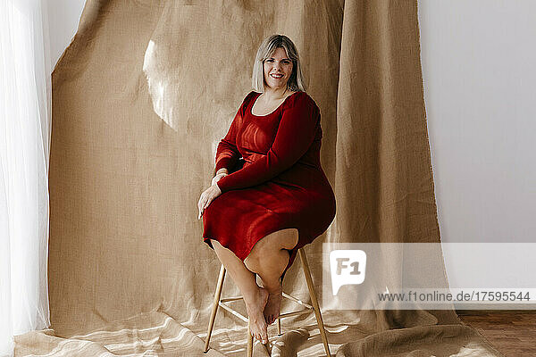 Smiling young curvy woman sitting on stool in front of brown background