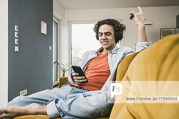 Happy man listening music through wireless headphones using smart phone on sofa at home