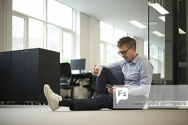 Businessman using smart phone sitting on ground in modern office