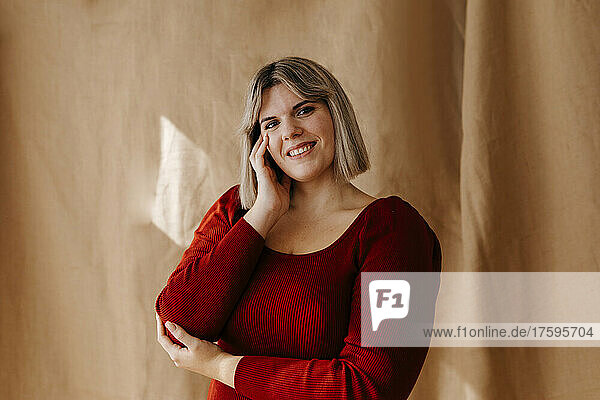 Happy beautiful curvy woman with hand on cheek standing in front of brown backdrop