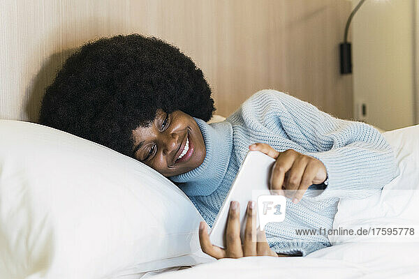 Smiling woman with turtleneck sweater watching on smart phone in bedroom at home